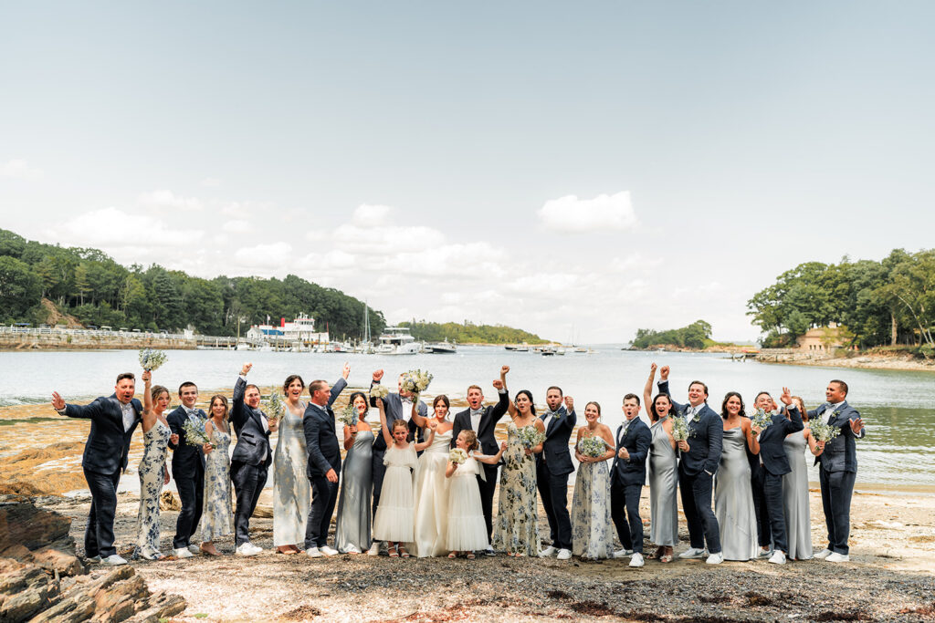 Wedding party cheering together on the shoreline with water and boats behind them at Inn at Diamond Cove in Maine.