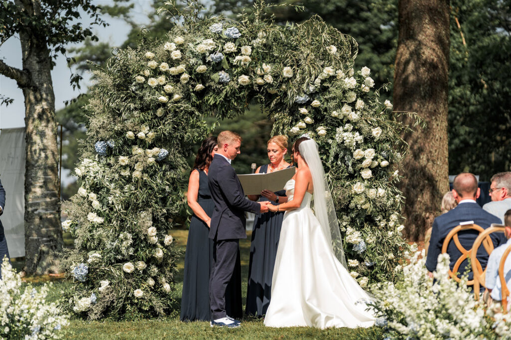 Bride and groom exchanging vows beneath a circular floral arch during an outdoor ceremony at Inn at Diamond Cove in Maine.