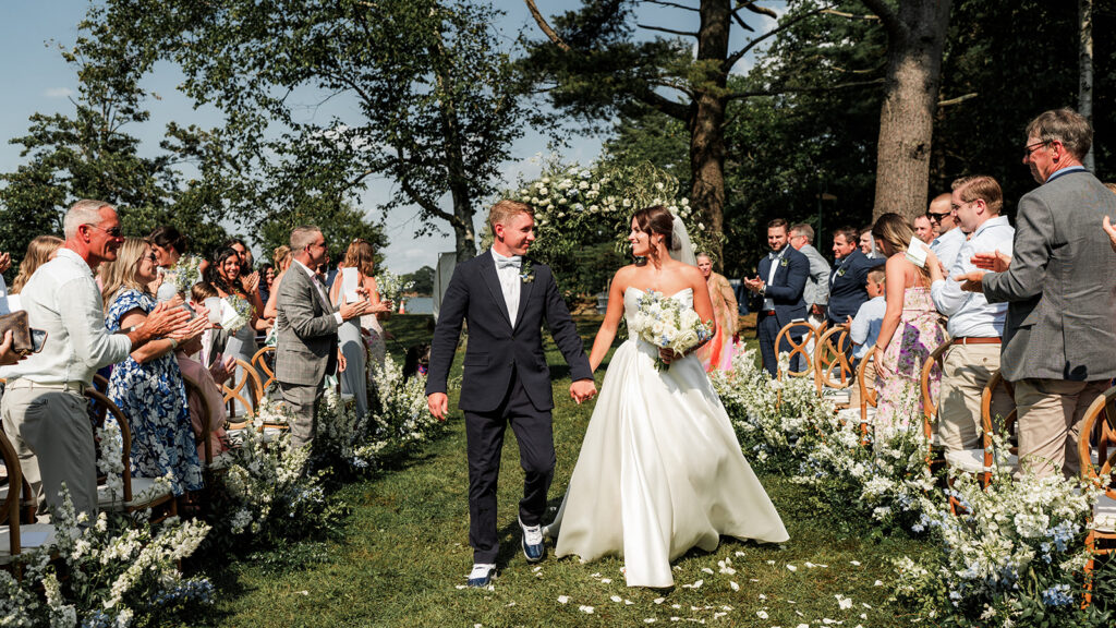 Bride and groom walk hand in hand down the aisle as guests stand and applaud at an outdoor ceremony at Inn at Diamond Cove in Maine.