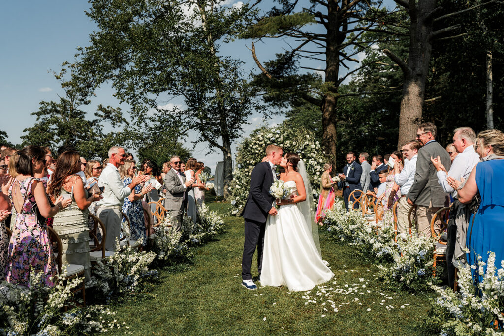 Bride and groom share a kiss at the end of the aisle surrounded by guests and white floral arrangements at Inn at Diamond Cove in Maine.