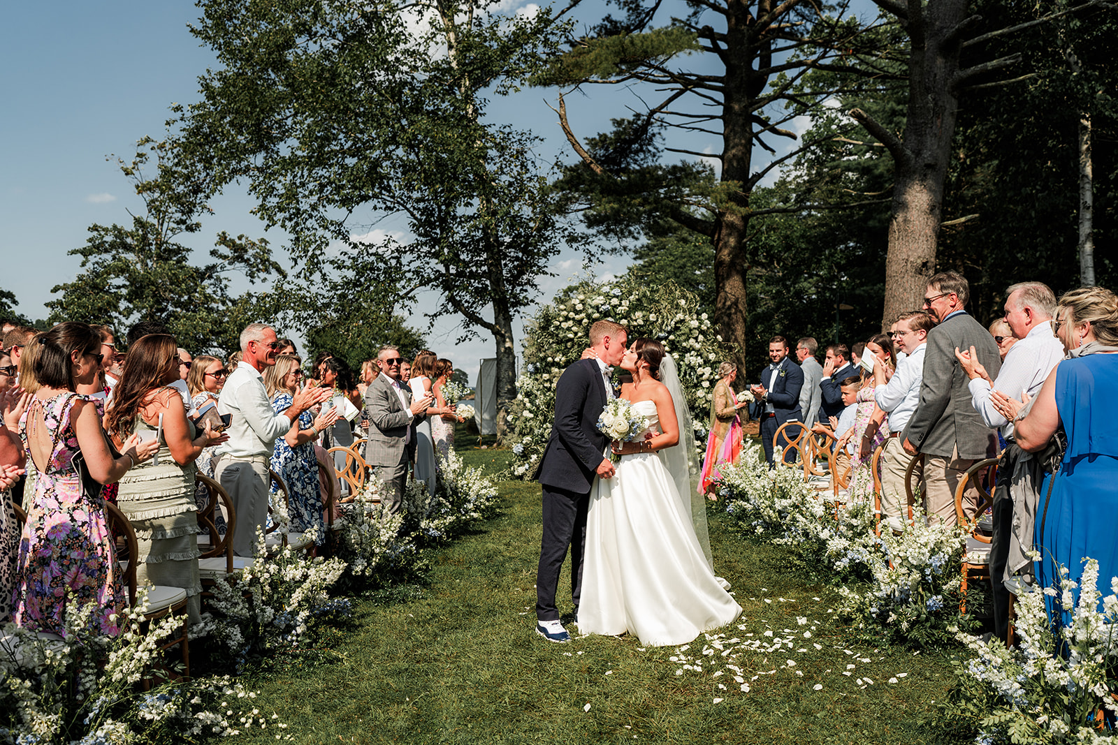 Bride and groom share a kiss at the end of the aisle surrounded by guests and white floral arrangements at Inn at Diamond Cove in Maine.