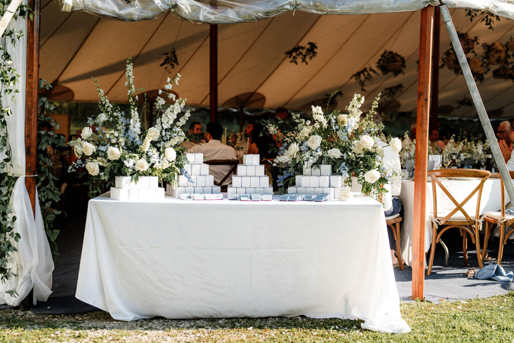 Escort card display with white and blue florals arranged on a table inside a reception tent.