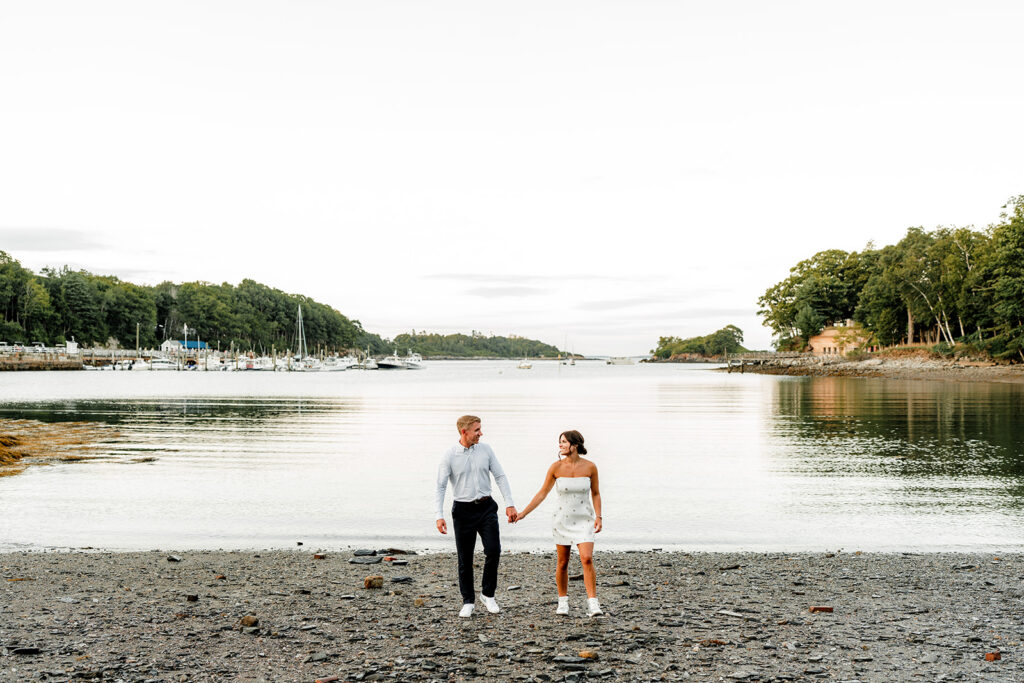 Bride and groom walking hand in hand along a rocky shoreline with boats and harbor views at Inn at Diamond Cove in Maine.