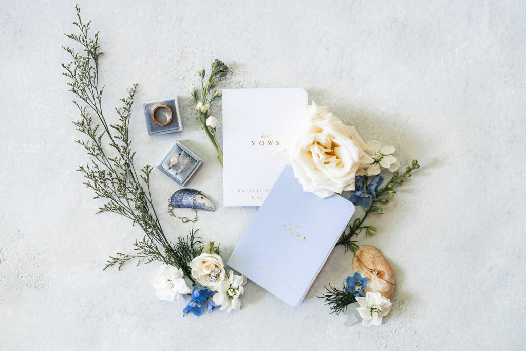 Wedding flat lay with vow books, rings, and soft blue and white florals styled on a light surface.