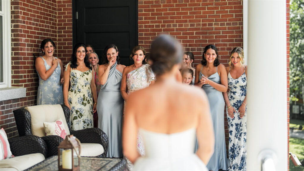 Bride seen from behind approaching a group of women reacting on a brick porch during wedding preparations at Inn at Diamond Cove in Maine.