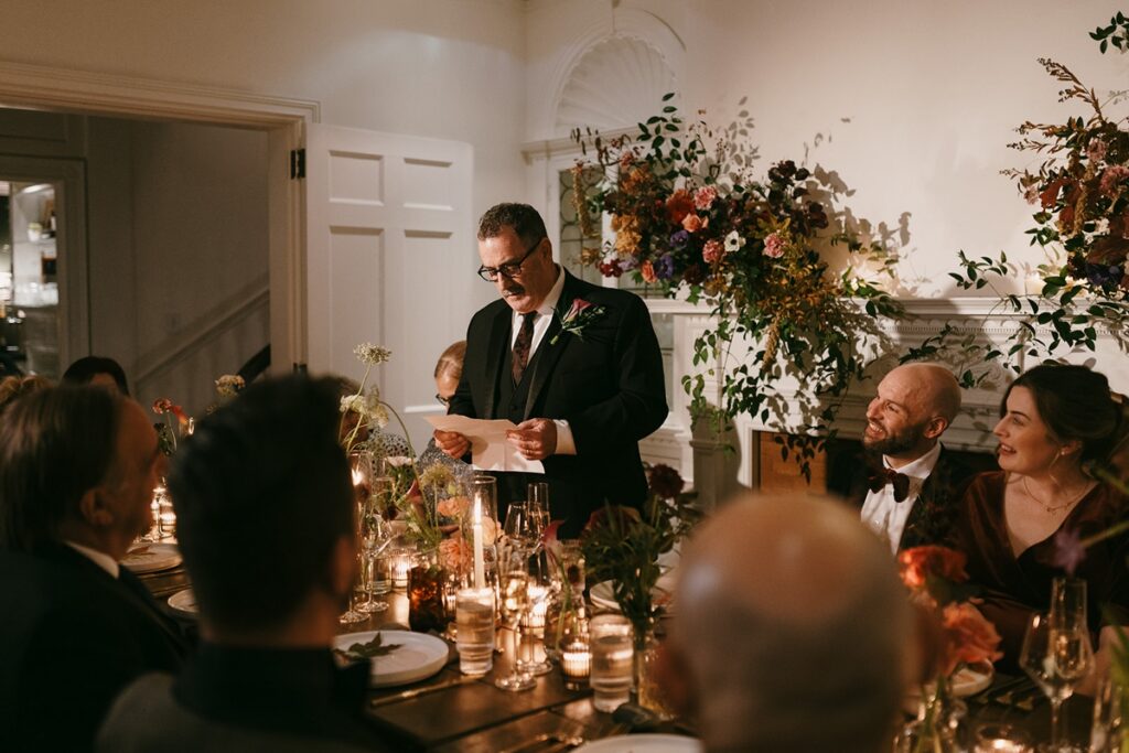 Guest giving a speech while holding paper at a candlelit reception table while guests look on smiling.
