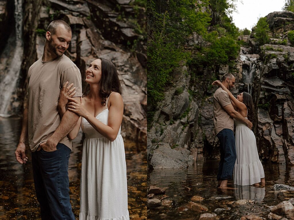 Couple standing together in a shallow stream beneath rocky cliffs and embracing barefoot below a New Hampshire waterfall during their engagement session.