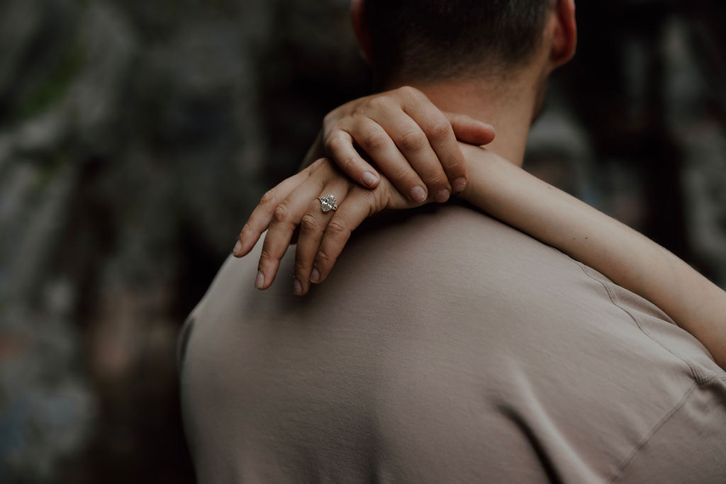 Close-up of engagement ring on woman's hand resting on partner’s shoulder during a waterfall engagement session.