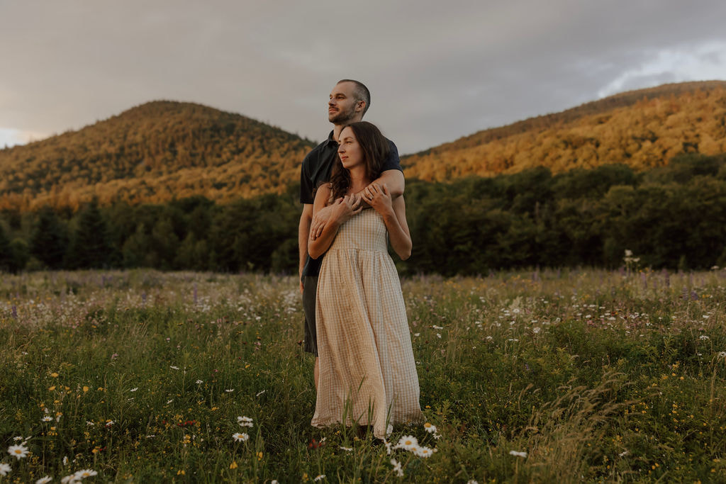 Couple standing together in a mountain wildflower meadow with forested hills behind them at sunset.