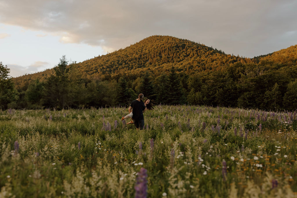 Couple in a wildflower meadow with a mountain backdrop at sunset as the man lifts the woman during a New Hampshire engagement session.