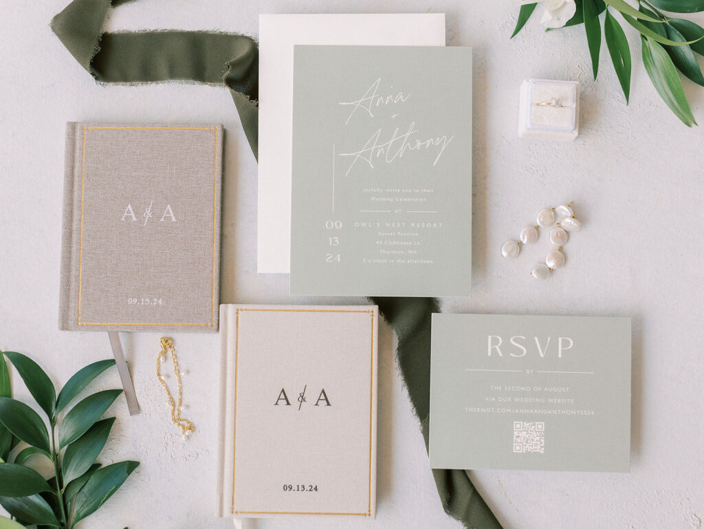 Flat lay of wedding invitation, vow books, rings, and RSVP card styled with greenery and olive ribbon.