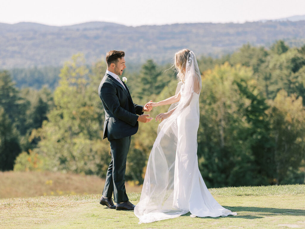 Bride and groom smiling and reaching for each other during their first look with mountain views behind them.