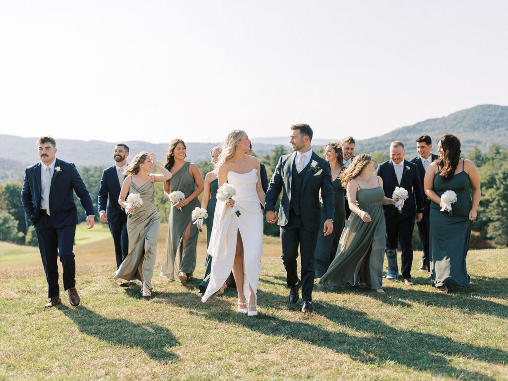 Bride and groom walking with bridesmaids and groomsmen across a grassy hill during wedding portraits.