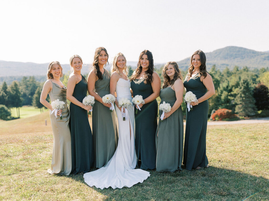 Bride surrounded by bridesmaids in green dresses holding white bouquets during outdoor wedding portraits.