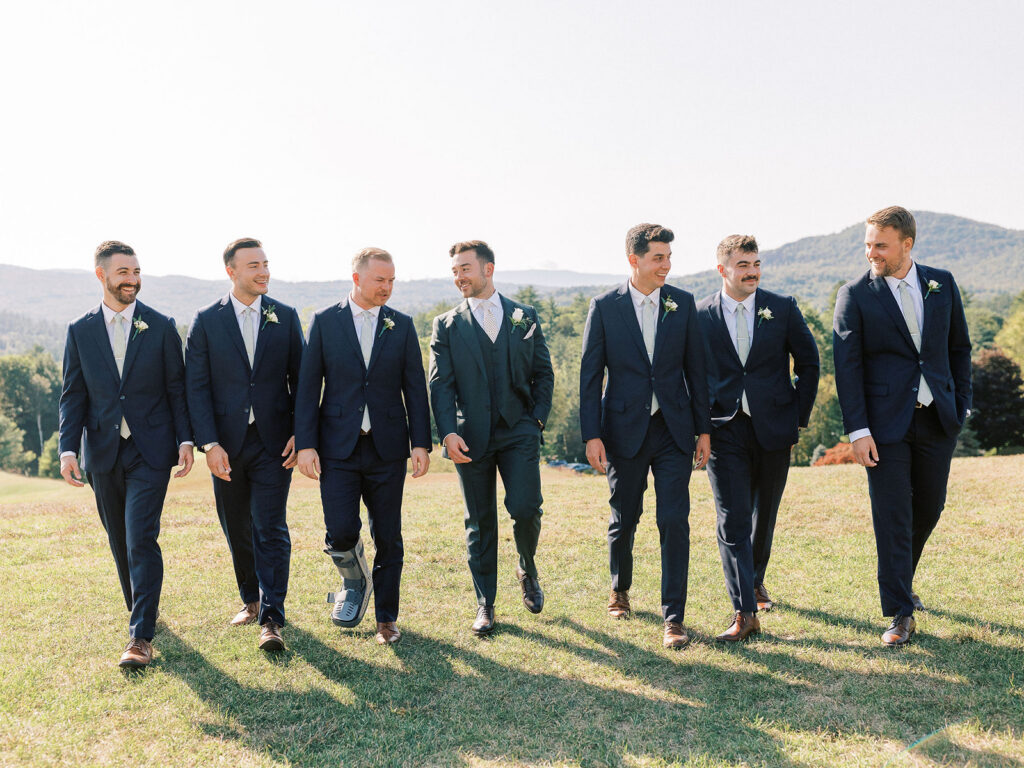 Groom walking with groomsmen in navy suits across a hill during wedding party portraits.