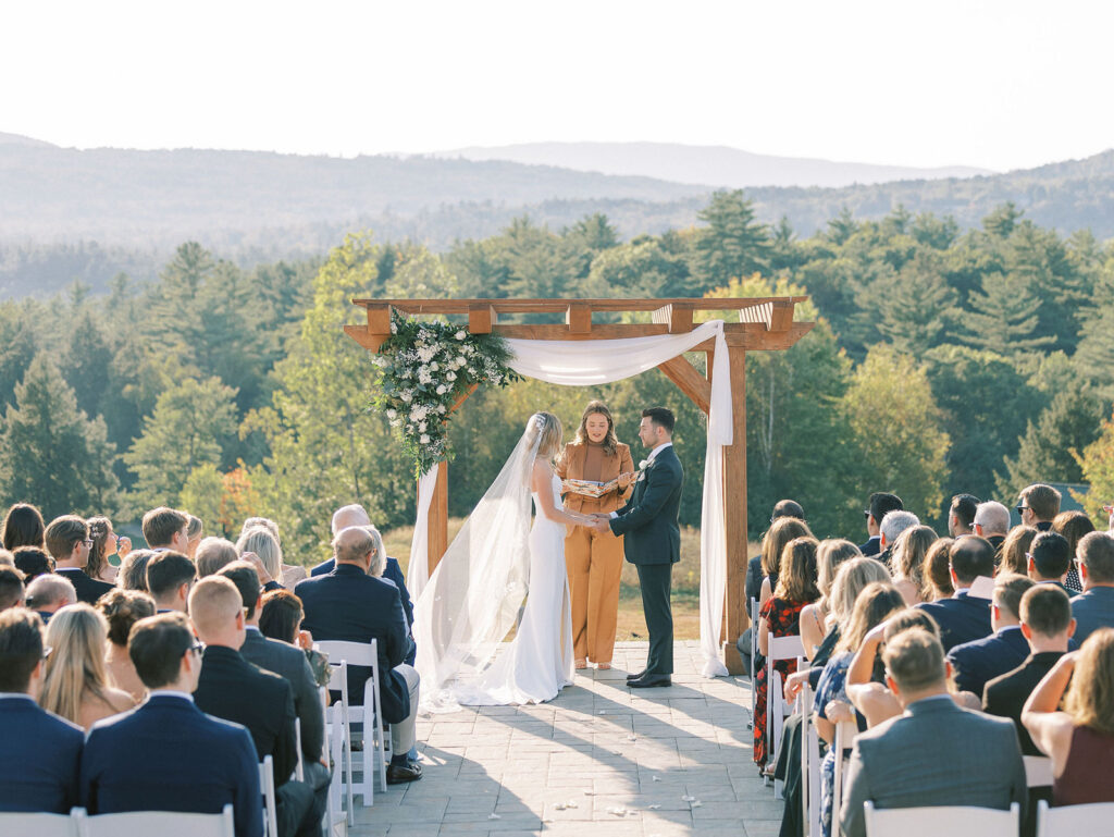 Bride and groom holding hands beneath a wooden arbor during their outdoor wedding ceremony with mountains behind them.