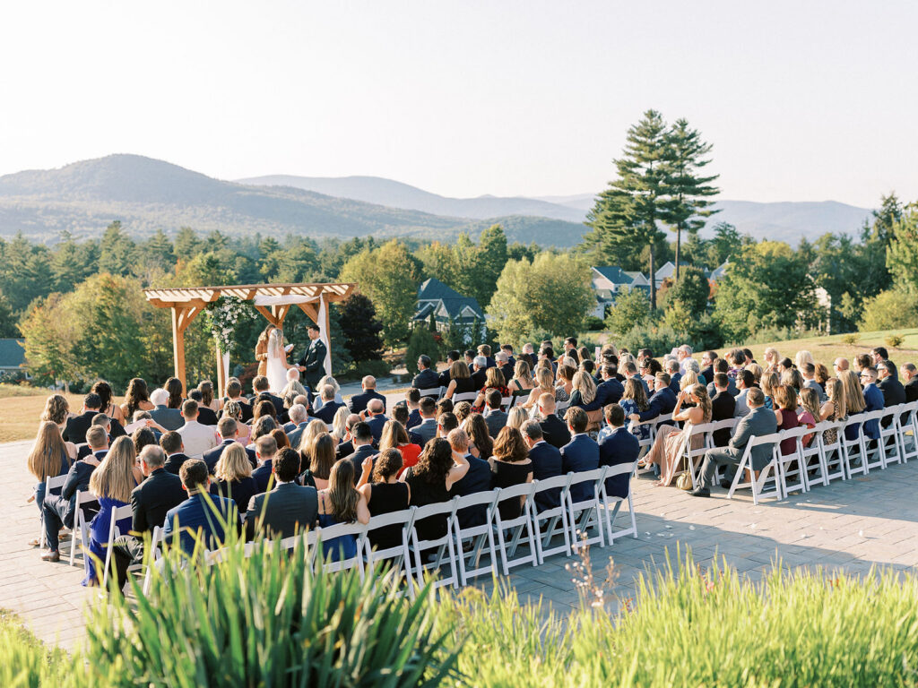 Guests seated during an outdoor ceremony as the couple exchanges vows beneath a wooden floral arbor.