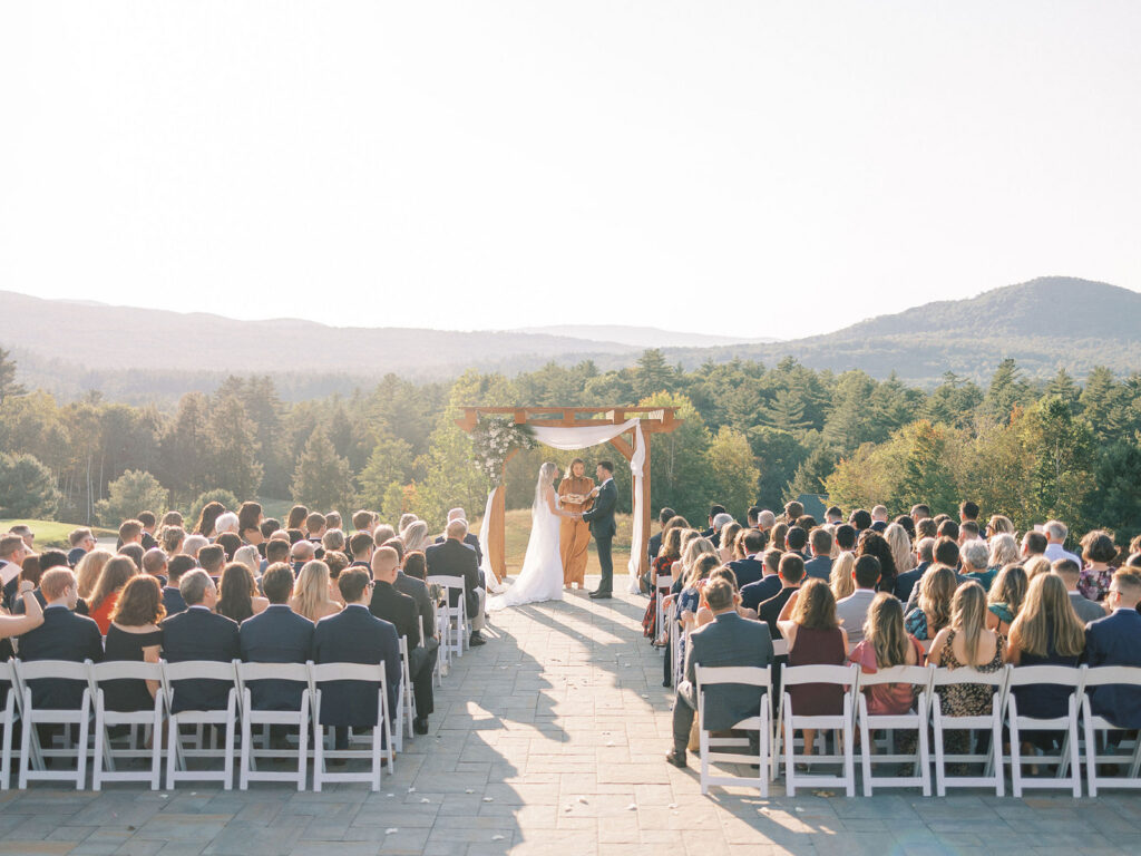 Wide view of bride and groom exchanging vows during a mountain-view ceremony surrounded by guests.