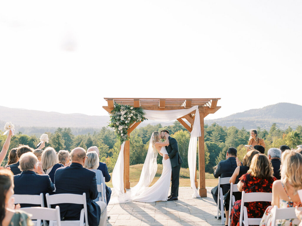 Bride and groom sharing their first kiss beneath a wooden arbor as guests celebrate during the ceremony.