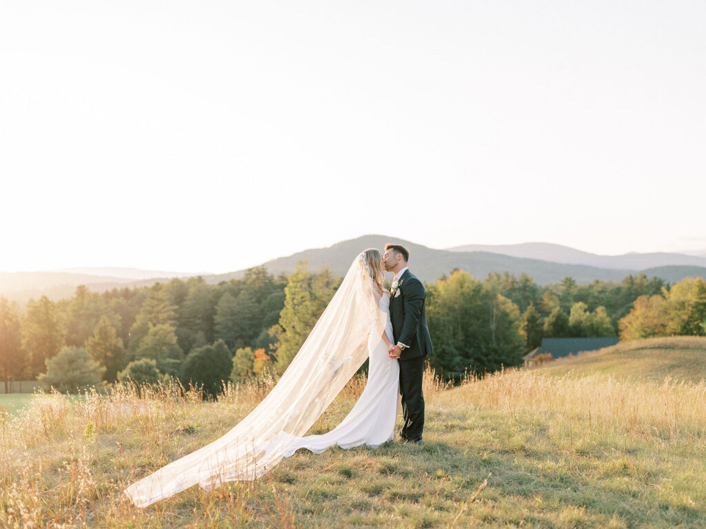 Bride and groom kissing during golden hour portraits with a flowing veil and mountain landscape behind them.