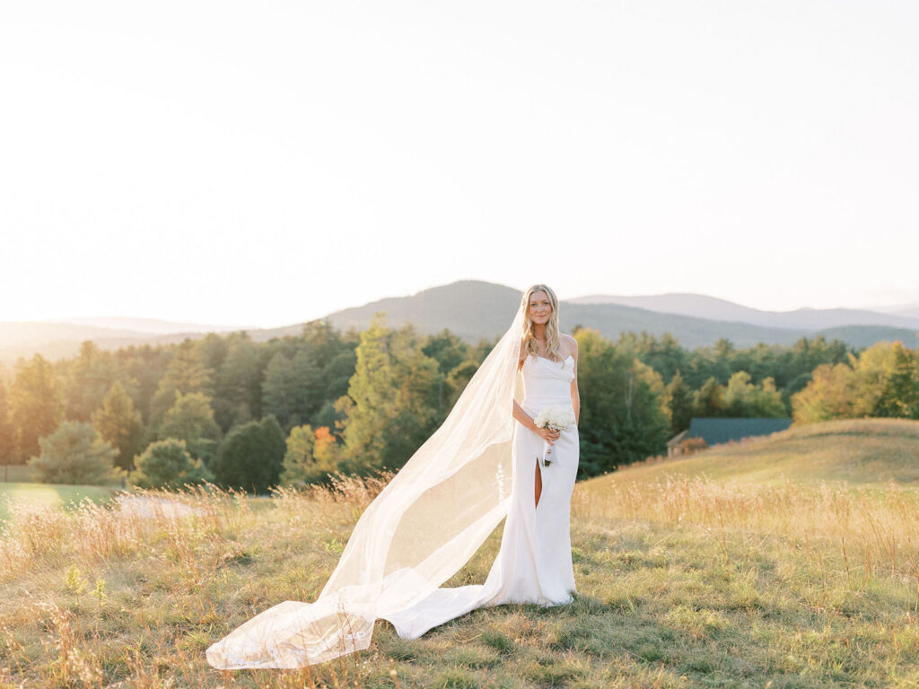 Bride standing in tall grass holding her bouquet as her long veil flows in the sunset light.