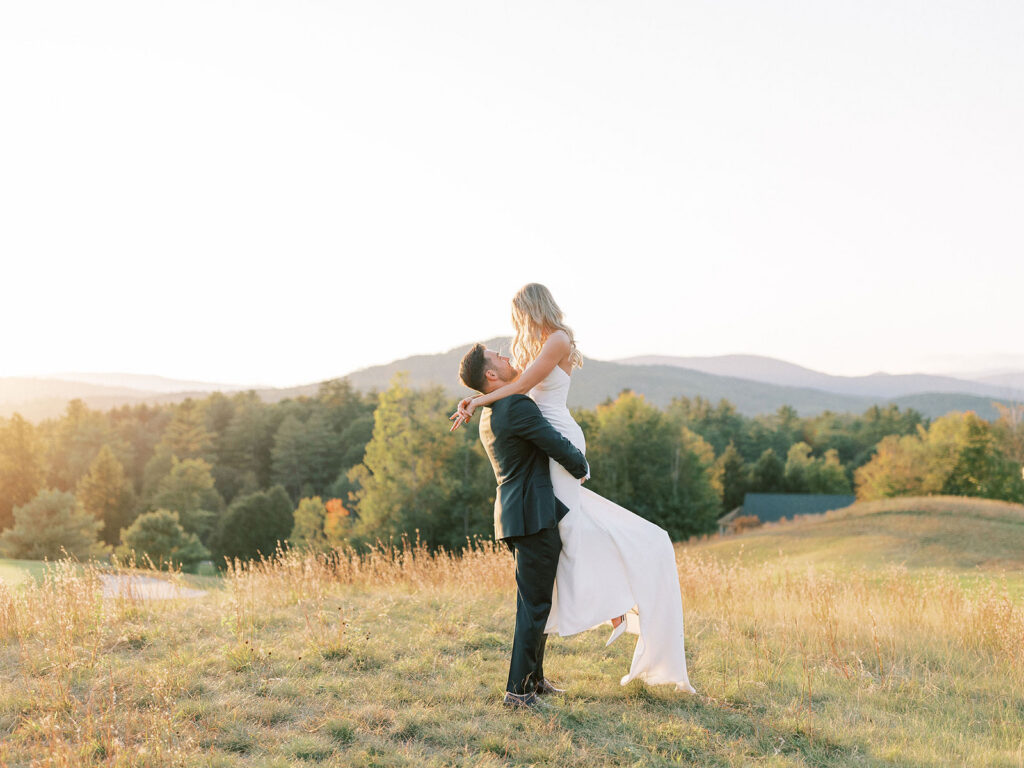 Groom lifting the bride in a grassy field during golden hour portraits with mountains in the background.
