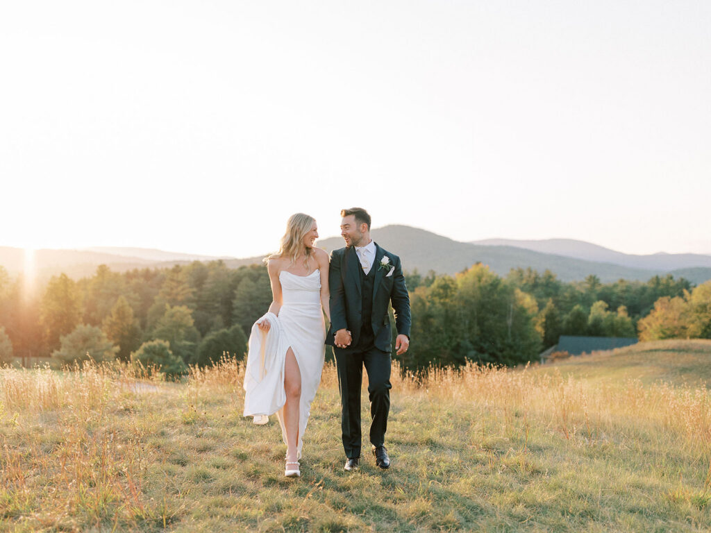 Bride and groom walking hand in hand through a field during sunset wedding portraits.