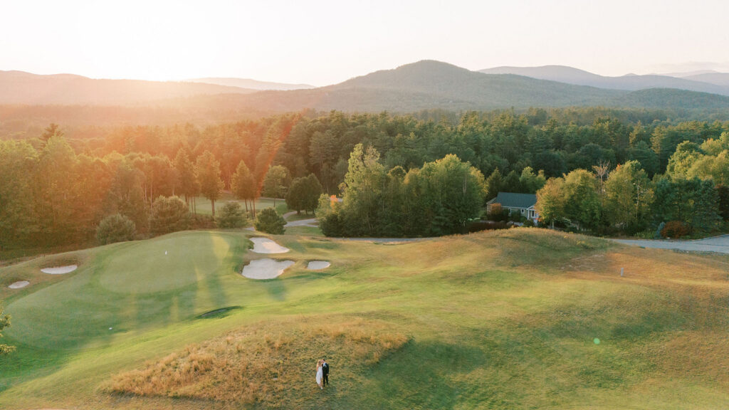 Aerial view of the couple standing on a hill surrounded by a golf course and mountain landscape at sunset.