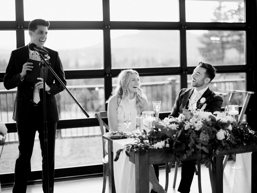 Wedding guest giving a speech while the bride and groom laugh at their reception table.
