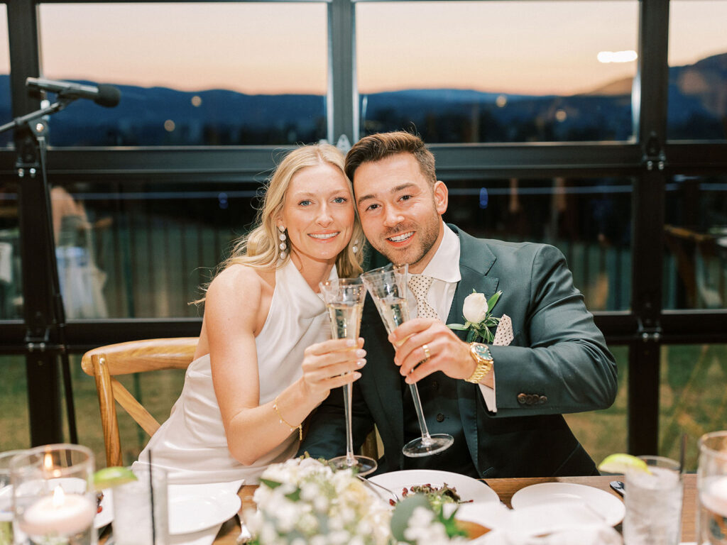 A bride and groom toasting with champagne flutes at their sweetheart table, mountain sunset visible through the windows behind them.