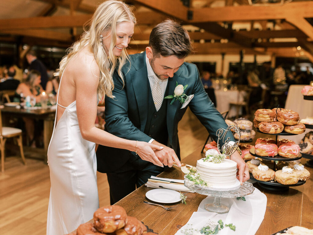 A bride and groom cutting a white single-tier wedding cake surrounded by tiered donut displays at a rustic reception.