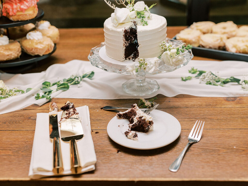 A sliced white-frosted chocolate wedding cake on a glass pedestal, flanked by donuts, with gold cake utensils in the foreground.