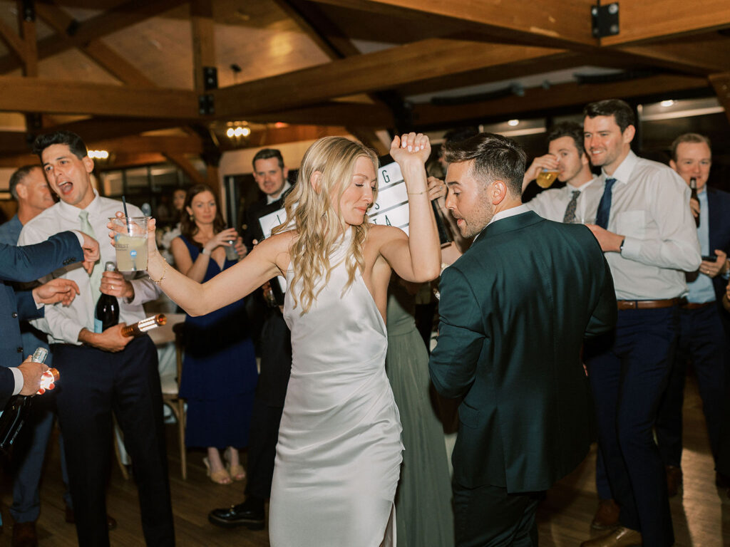 The bride in a white satin gown dancing with the groom in a navy suit, surrounded by cheering guests in a timber-framed venue.