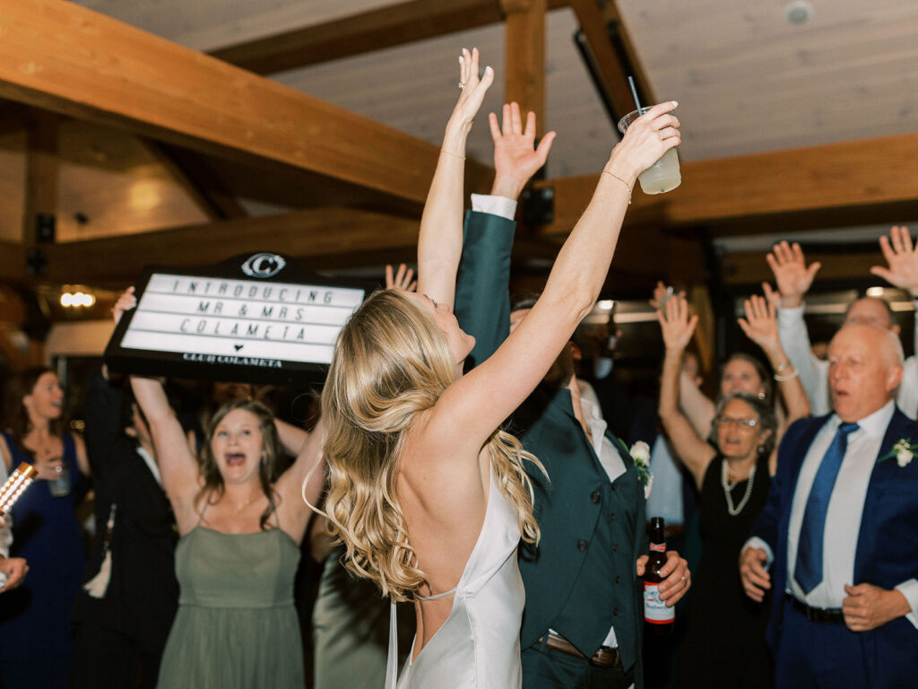 The bride and groom celebrating with arms raised as guests cheer behind a sign reading "Introducing Mr & Mrs Colameta."