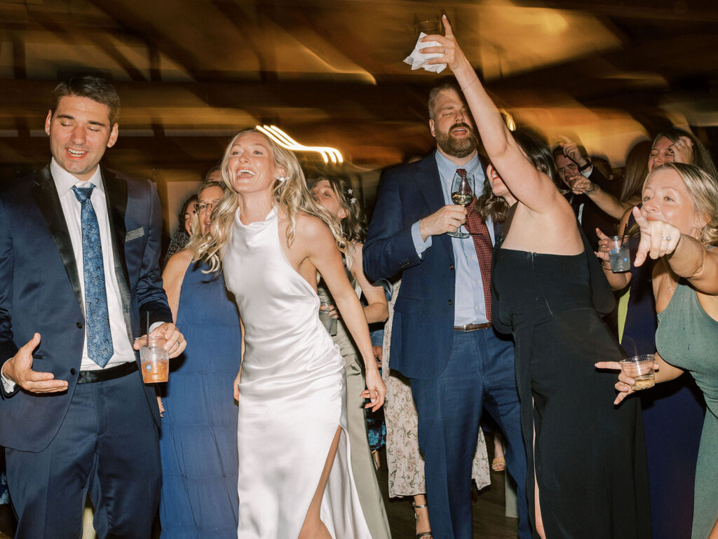 The bride laughing and dancing with guests holding drinks on a lively reception dance floor.