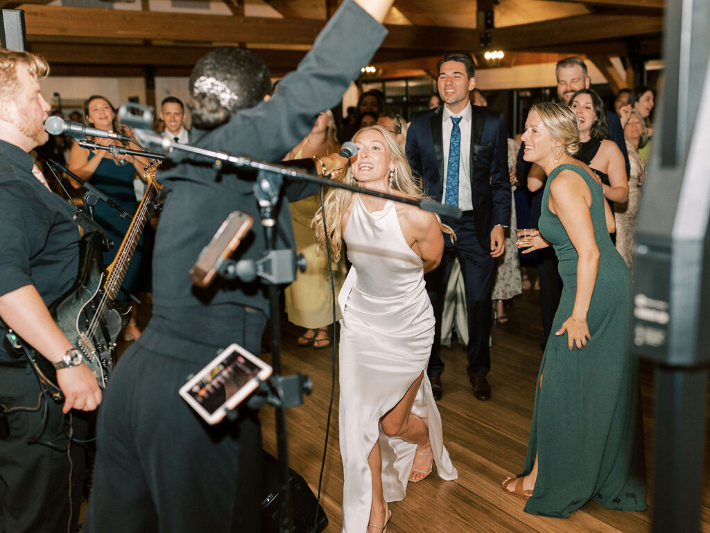 The bride singing into a microphone at the front of the live band stage, delighting guests behind her.