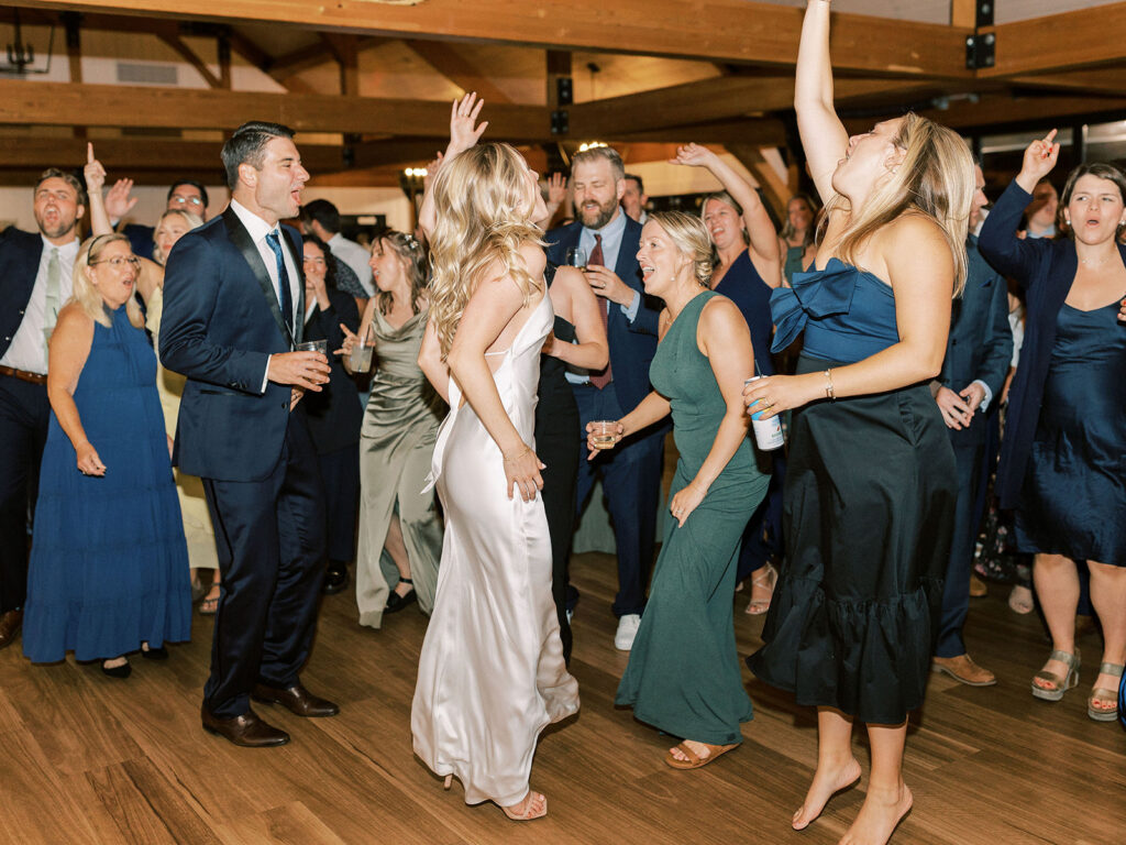The bride in a white satin gown dancing with a crowd of energetic guests in a wood-beamed reception hall.