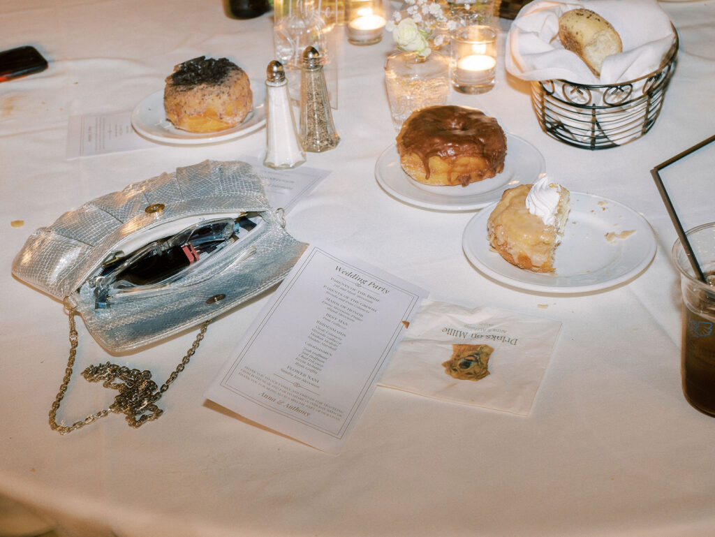 A candid reception table scene with donuts, a wedding program, an open silver clutch, and a cocktail napkin reading "Drinks on Millie."
