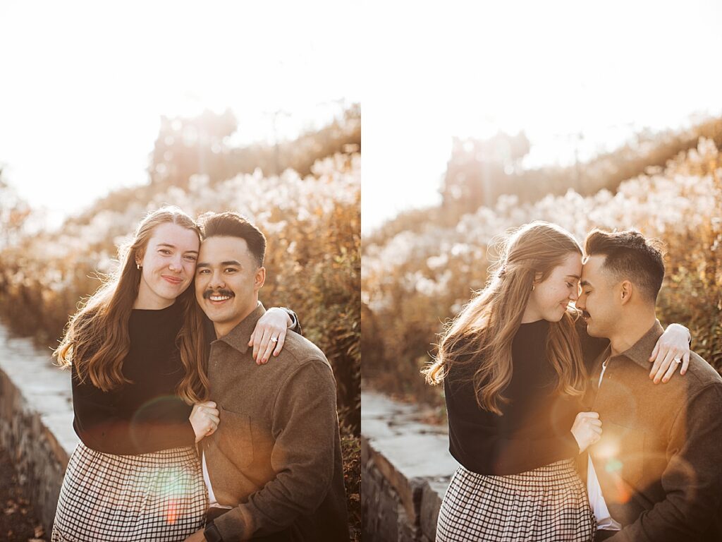 Engaged couple sitting along stone path with sun flare during golden hour, smiling and leaning together during coastal engagement session.