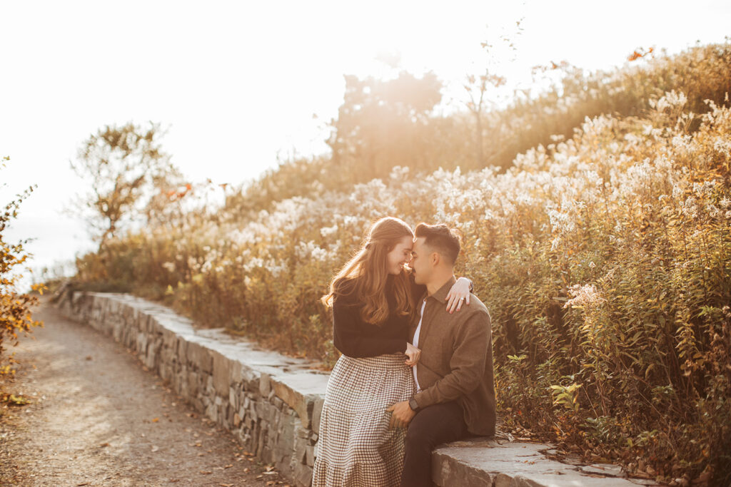 Engaged couple sitting together on stone wall along coastal path surrounded by tall grasses during warm sunset light.