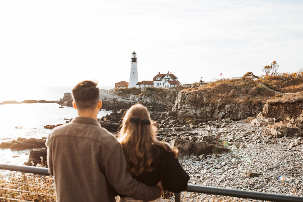 Couple stands arm in arm looking out toward Portland Head Light lighthouse and rocky shoreline at sunset.