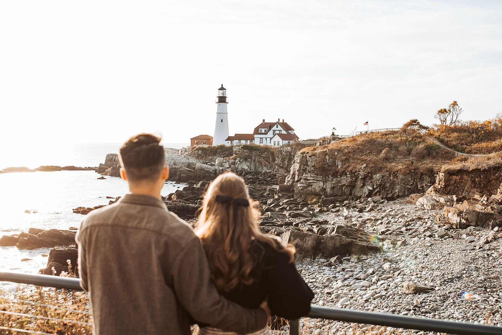 Couple stands arm in arm looking out toward Portland Head Light lighthouse and rocky shoreline at sunset.
