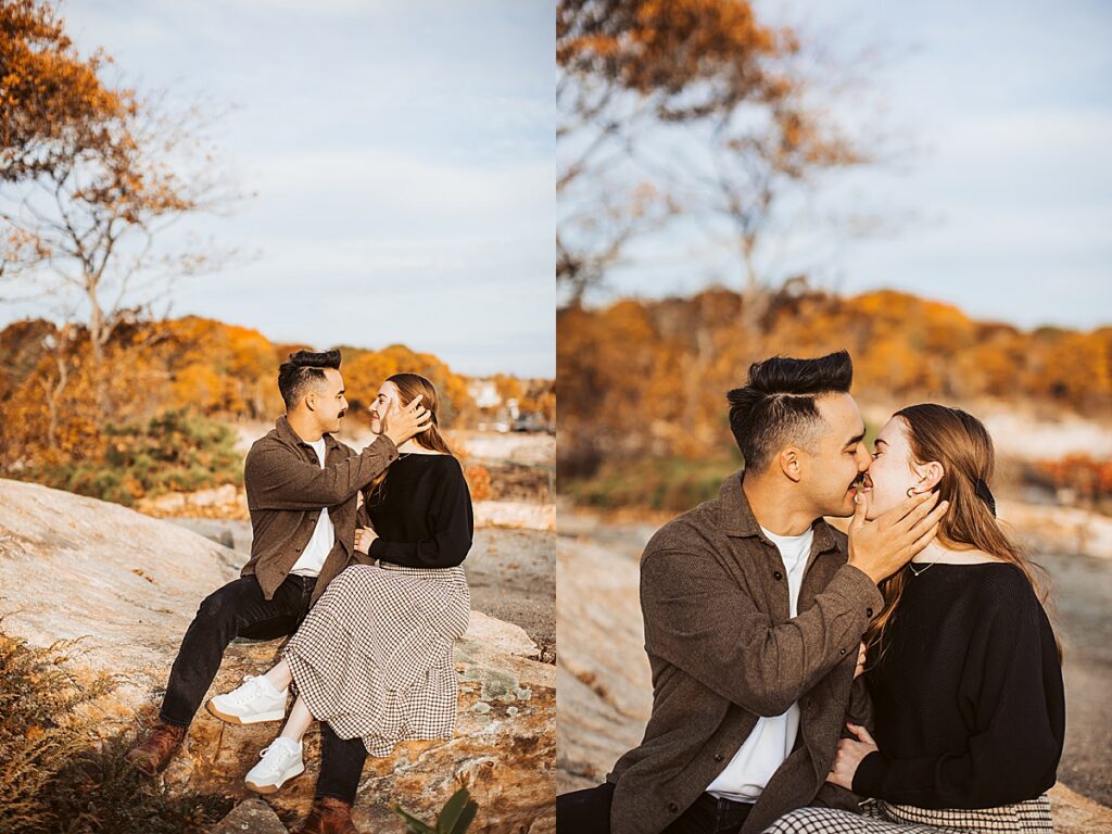 Engaged couple sitting on seaside rocks with autumn foliage, sharing a kiss during warm golden hour engagement photos.