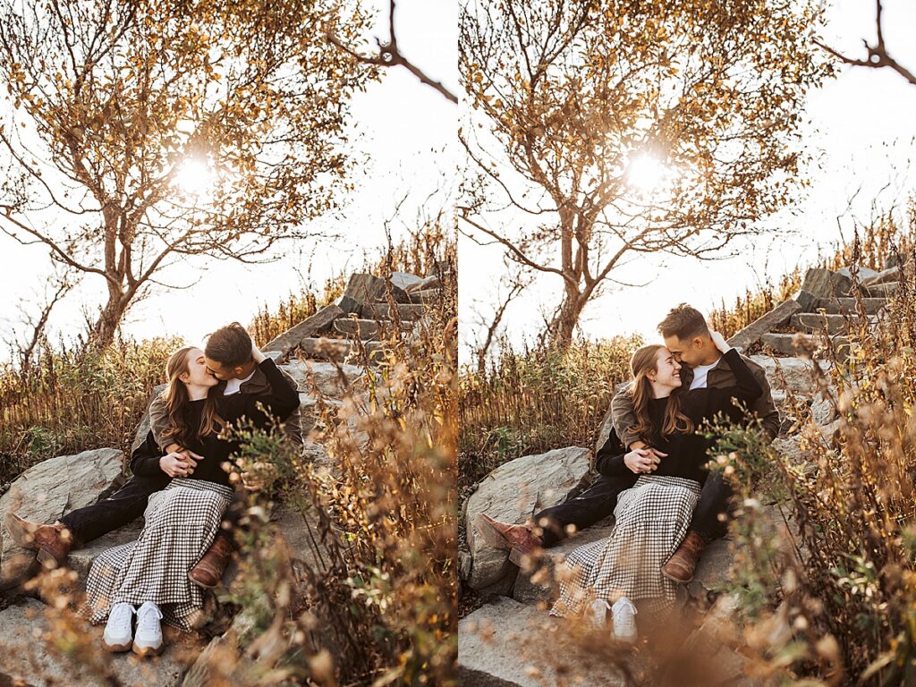 Couple cuddles and kisses while seated among coastal grasses and rocks with glowing sunset light filtering through trees.