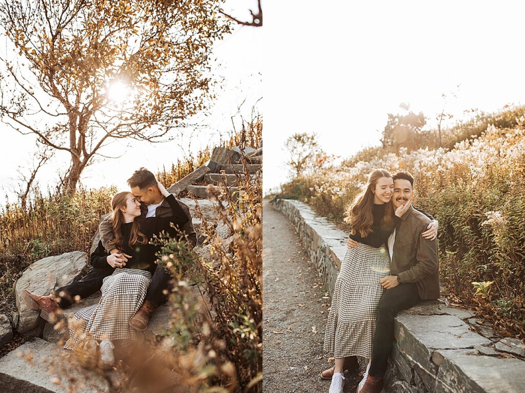 Engaged couple sitting along stone path surrounded by tall grasses during golden hour coastal engagement session.