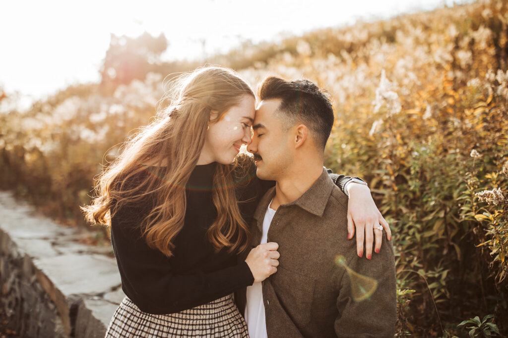 Couple standing close with foreheads touching along coastal path during golden hour engagement session with warm sunset light.