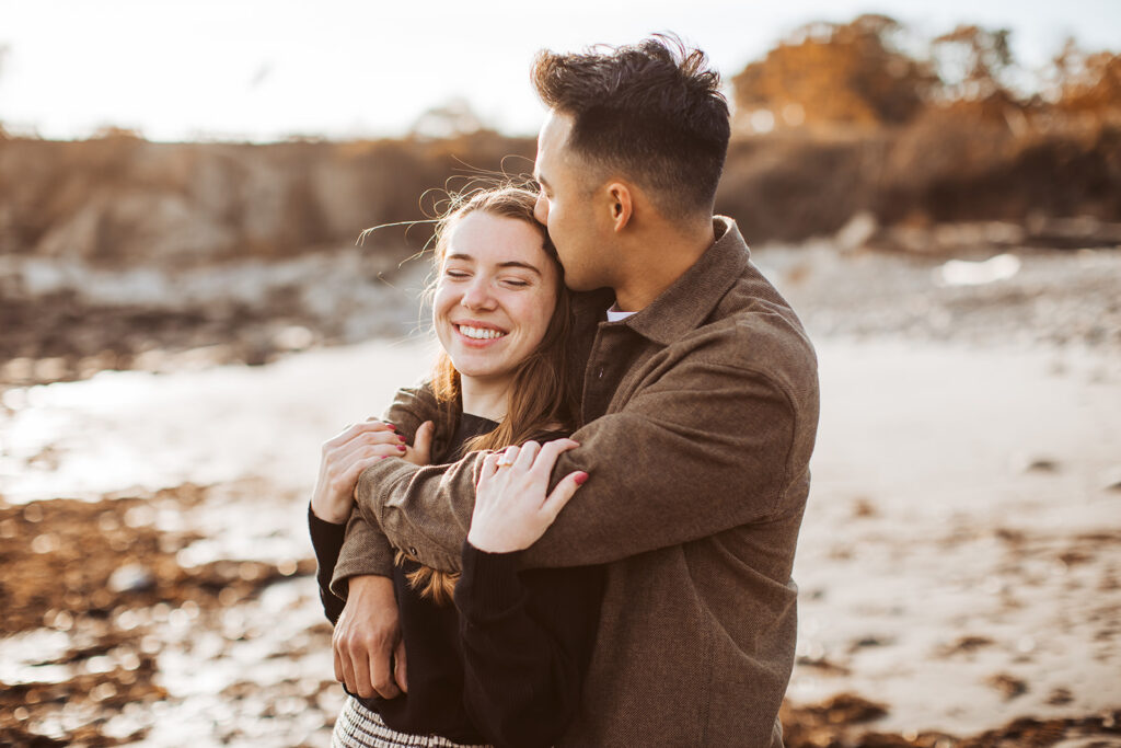 Man wraps arms around partner from behind and kisses her head as they stand together on rocky beach at sunset.