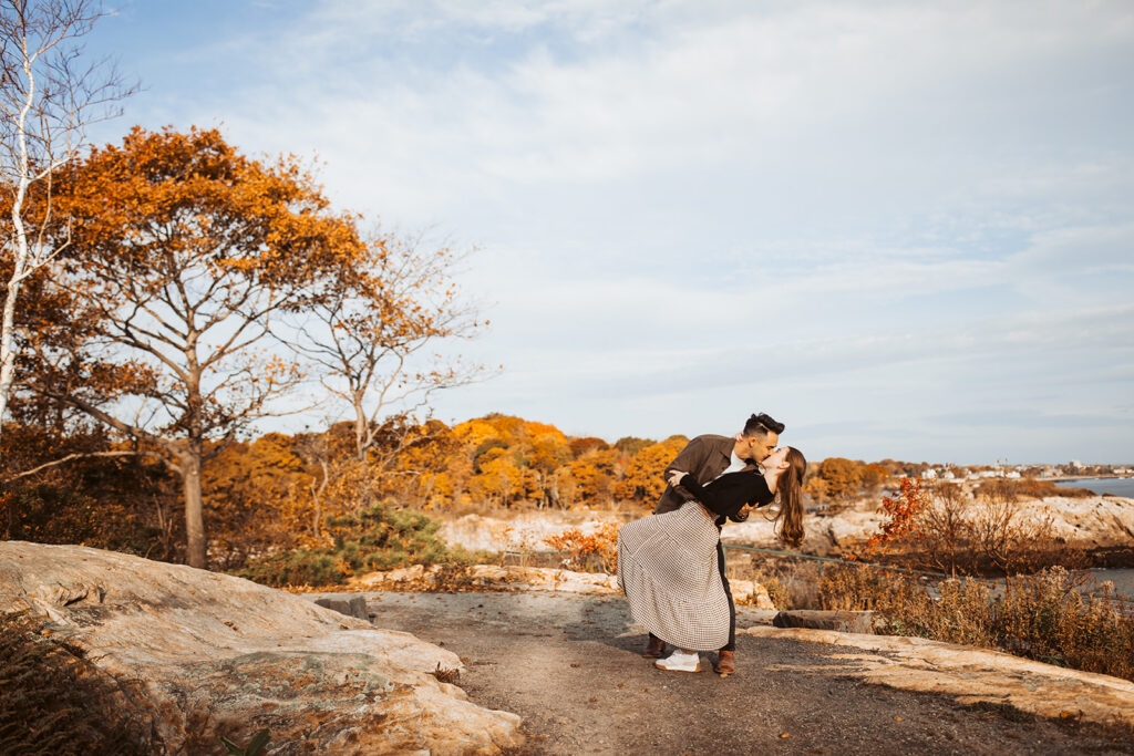 Couple shares a kiss while he dips her on scenic coastal overlook with autumn trees and rocky shoreline in background.