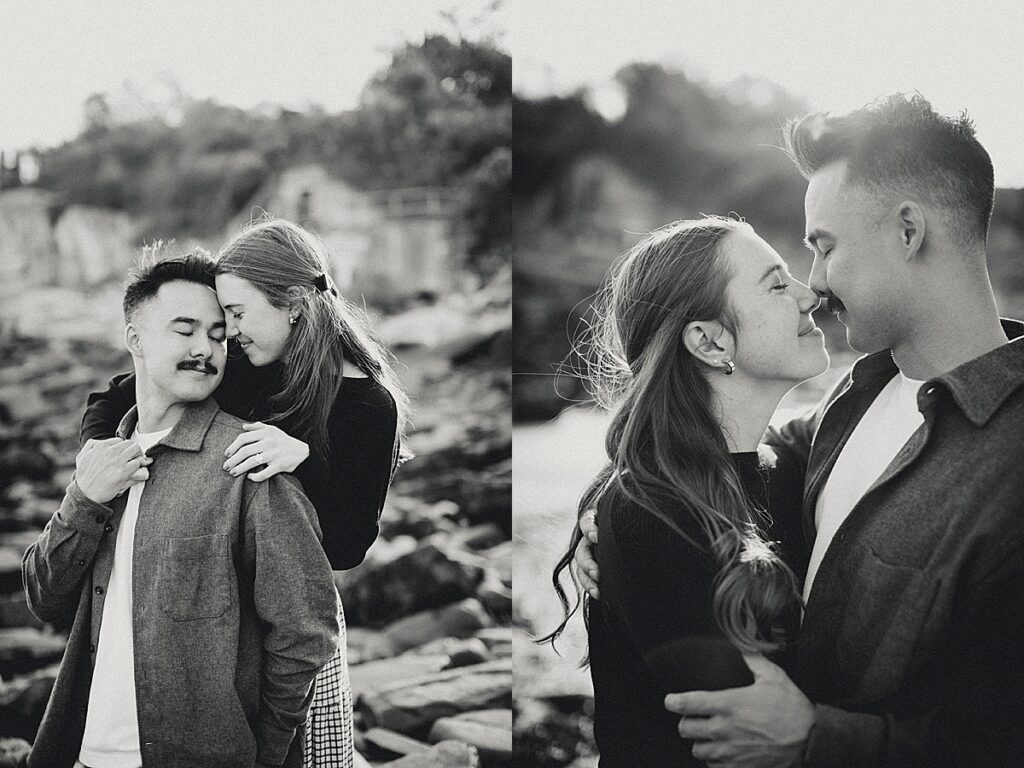 Black and white engagement portraits of couple embracing and smiling together on rocky beach shoreline.