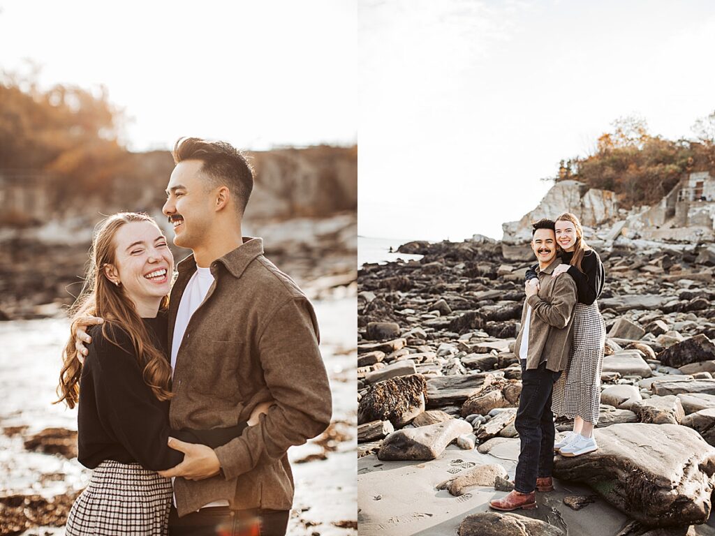 Couple laughing together and standing on rocky beach formations along Maine coastline during engagement session.